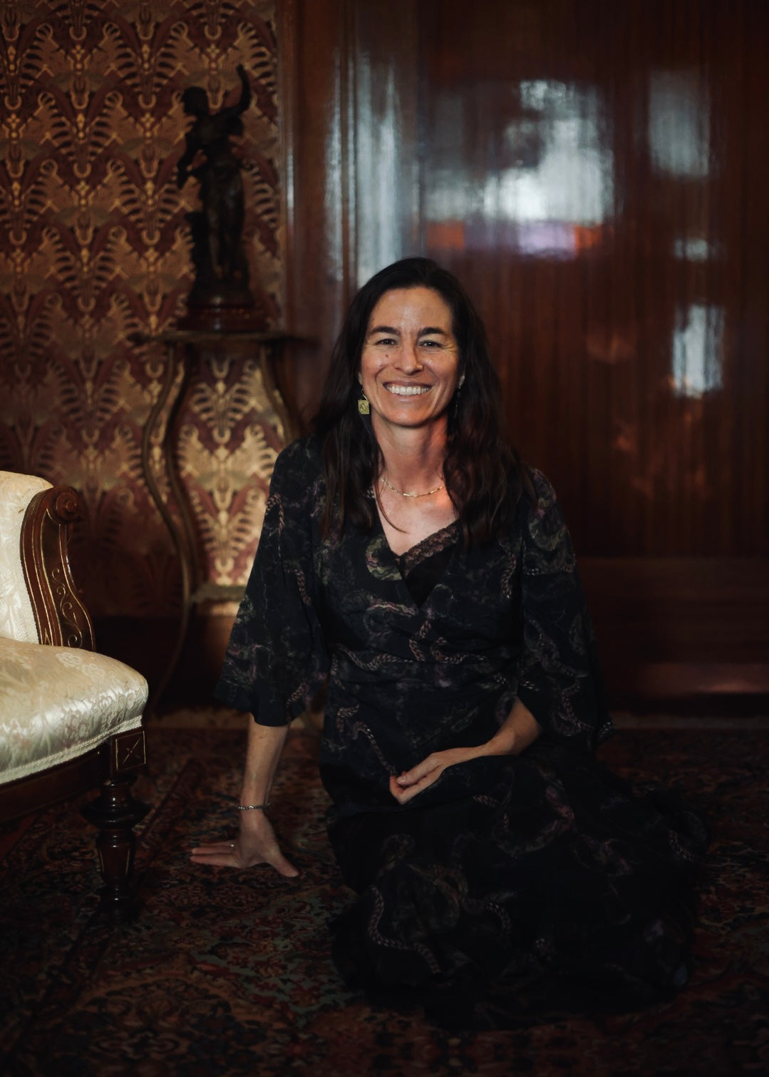 Woman in a dark dress sitting in an ornate room with patterned wallpaper and a statue.