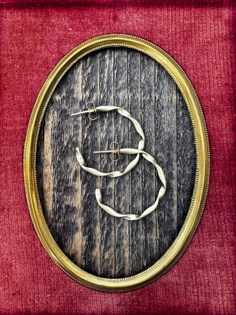 Twisted brass hoop earrings on a wooden surface within an oval frame, set against a red velvet background.