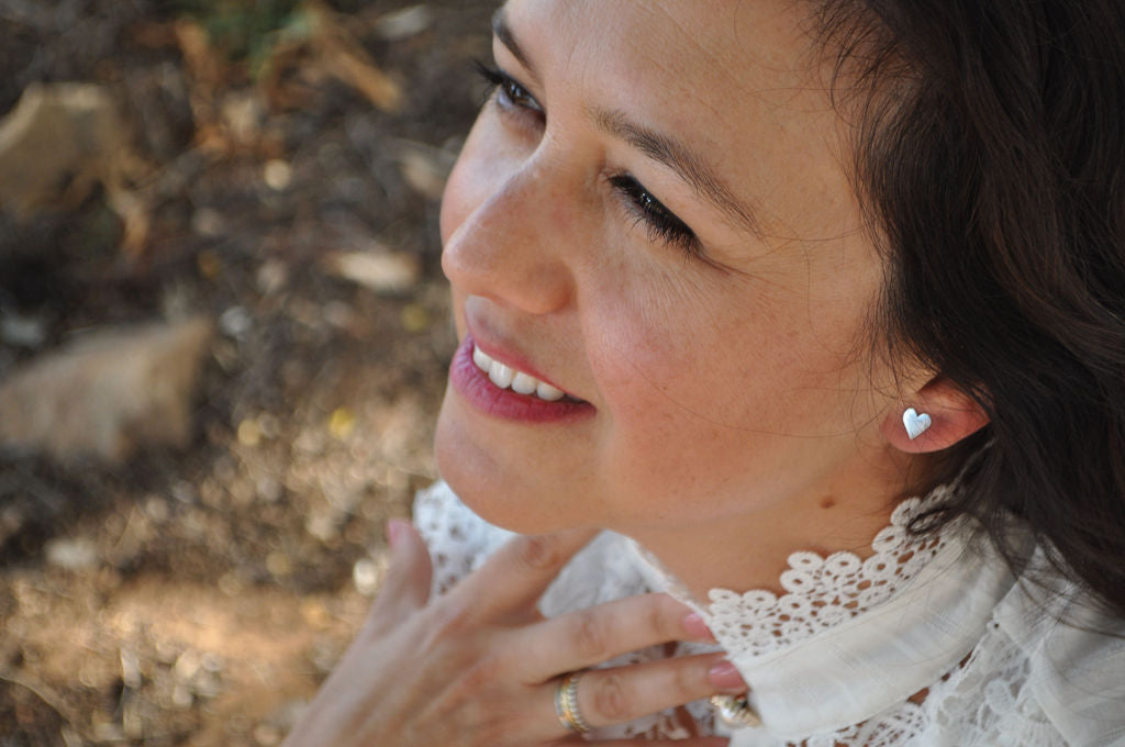 Model wearing heart-shaped Pharo sterling silver earring, looking upward while dressed in a white crochet top, standing in front of a whitewashed building.