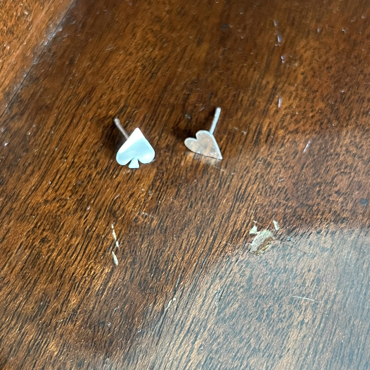 Close-up of Pharo sterling silver stud earrings shaped like a spade and heart, resting on a textured wooden surface, showcasing their sleek, polished design.